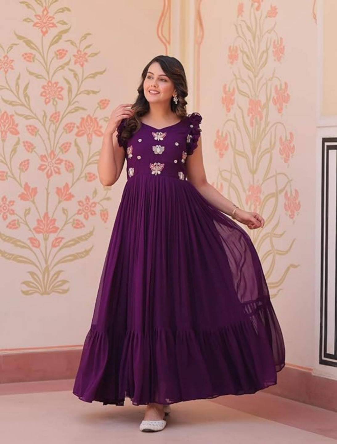 Woman wearing a purple dress with floral patterns against a decorative wall.