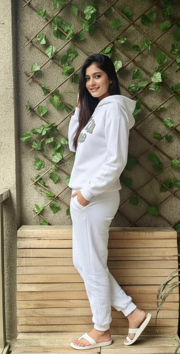 Woman in a white 'Lost in Space' hoodie and pants leaning against a wooden bench with greenery in the background.