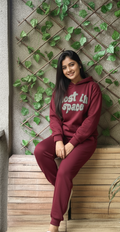 Woman wearing a maroon hoodie and pants with text, sitting against a wall with greenery.