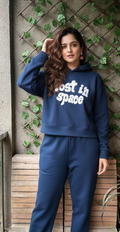 Woman wearing a blue hoodie with 'Lost in Space' text against a textured wall with greenery.