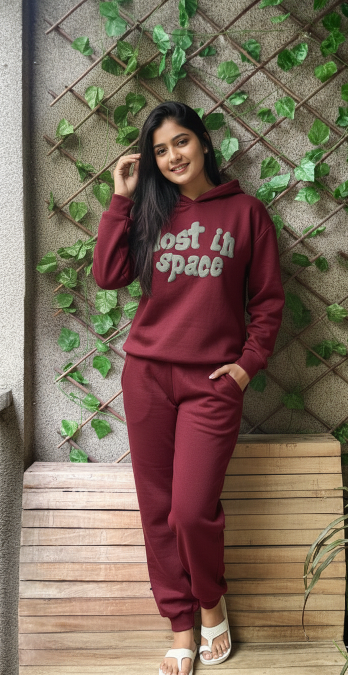 Woman wearing a maroon hoodie and pants with text, sitting against a wall with greenery.