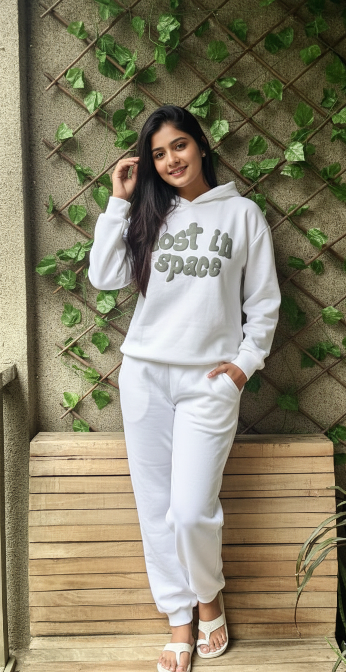 Woman in a white 'Lost in Space' hoodie and pants leaning against a wooden bench with greenery in the background.
