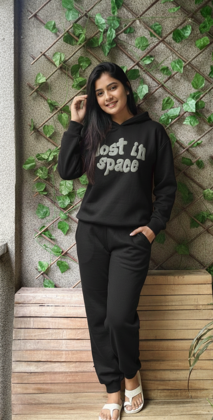 Woman wearing a black hoodie with 'Lost in Space' text, standing against a wall with greenery.