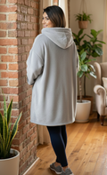 Woman wearing a gray hoodie in a cozy living room with plants and a brick wall.