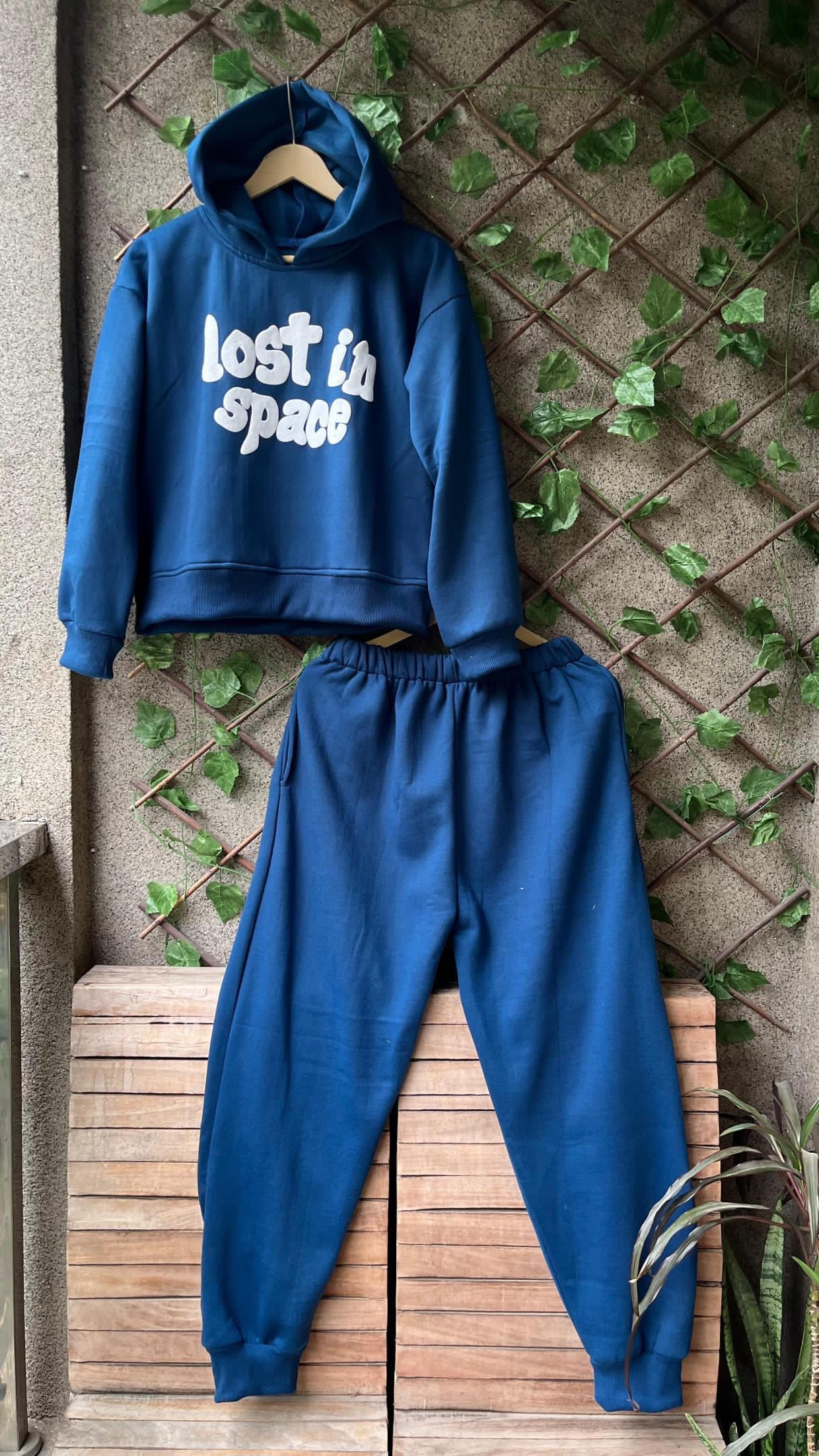 Blue hoodie and pants set with 'lost in space' text on a wooden surface with green leaves.