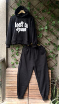 Black hoodie and pants set with 'Lost In Space' text on a wooden bench against a textured wall.