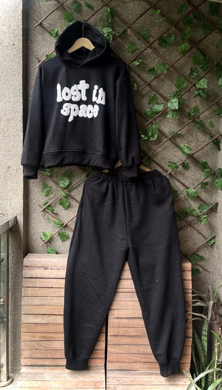 Black hoodie and pants set with 'Lost In Space' text on a wooden bench against a textured wall.
