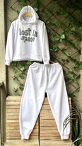 White hoodie and pants set with 'lost in space' text on a wooden bench against a leafy wall.