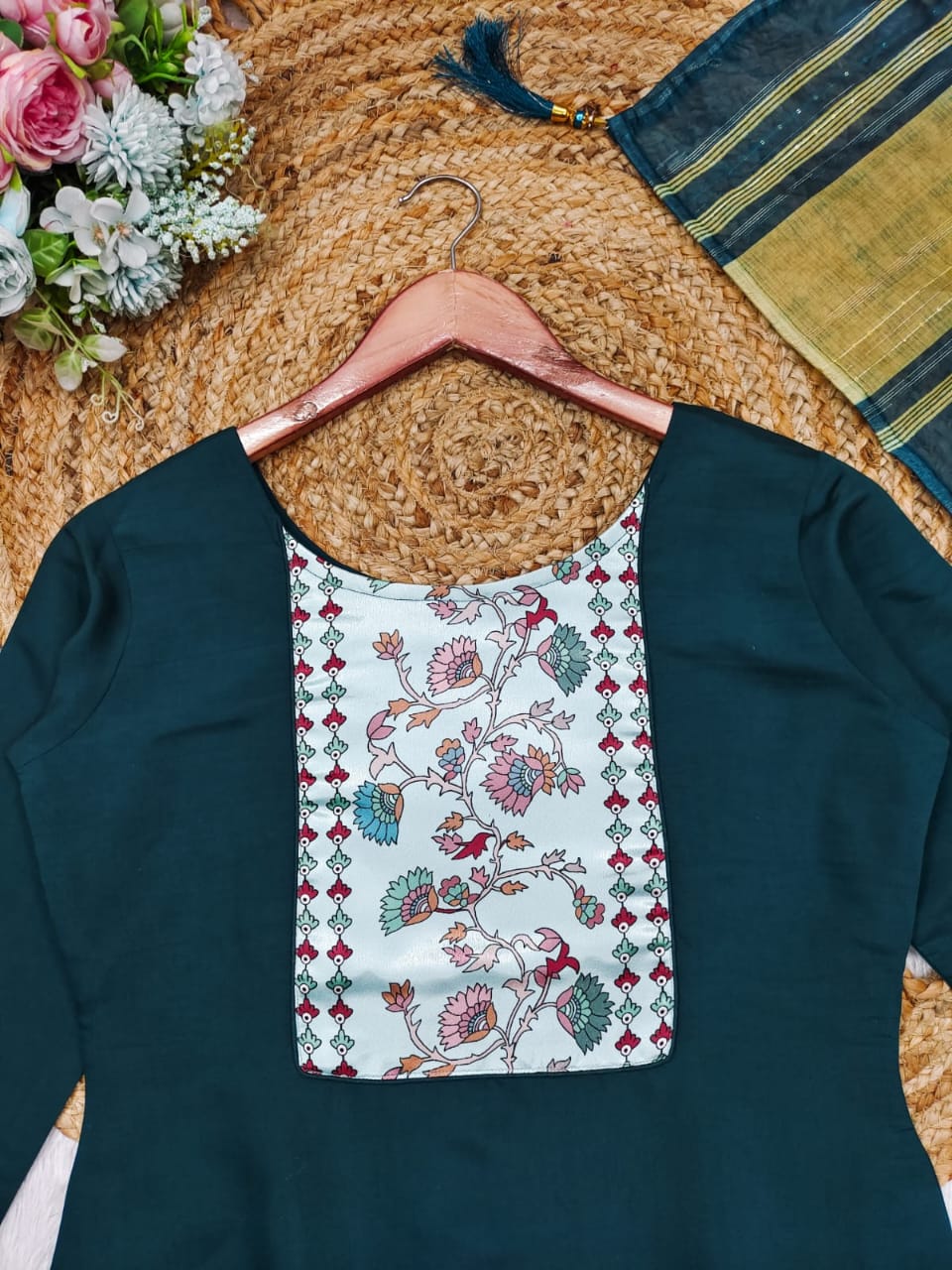 Green dress with floral pocket on a hanger, surrounded by flowers and a woven mat.