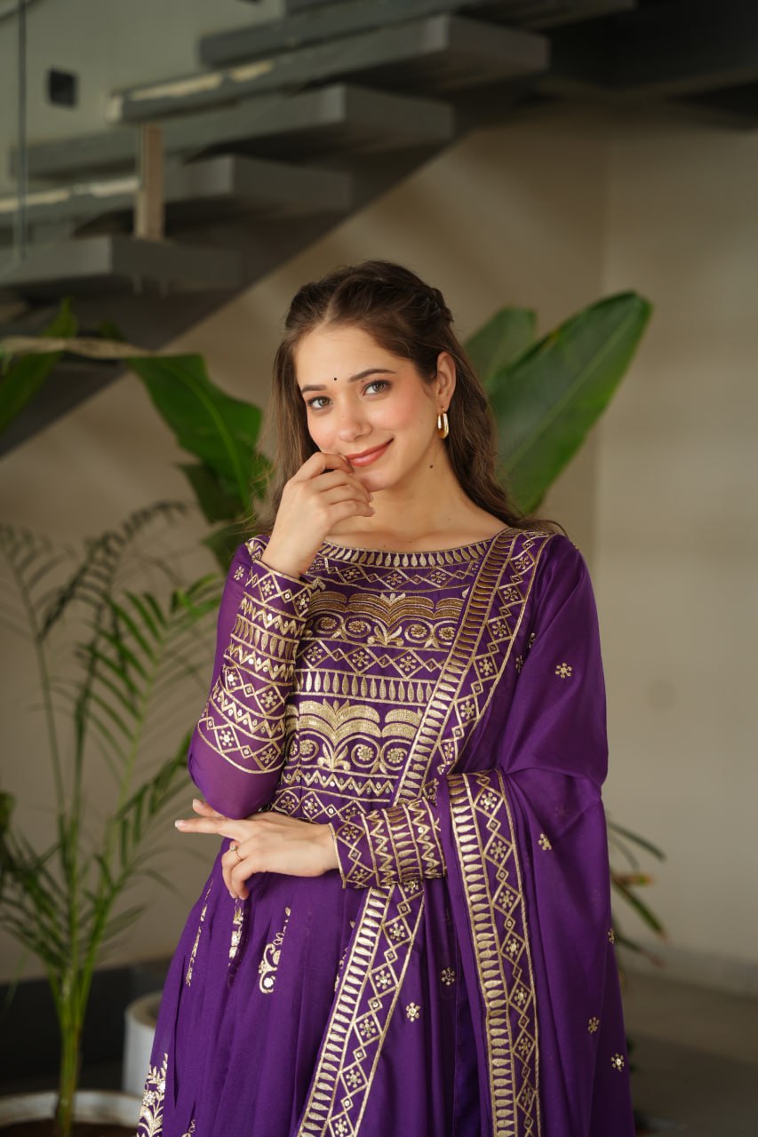 Woman in a purple traditional outfit standing indoors with plants and a staircase in the background